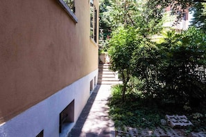 A narrow pathway flanked by a building with a beige facade and a small garden area with steps leading to a door.