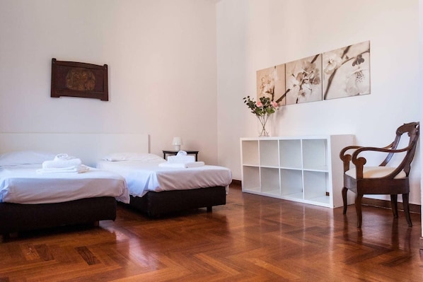 A hotel room with two beds, a wooden floor, a white console table, a chair, and a wall with floral artwork.