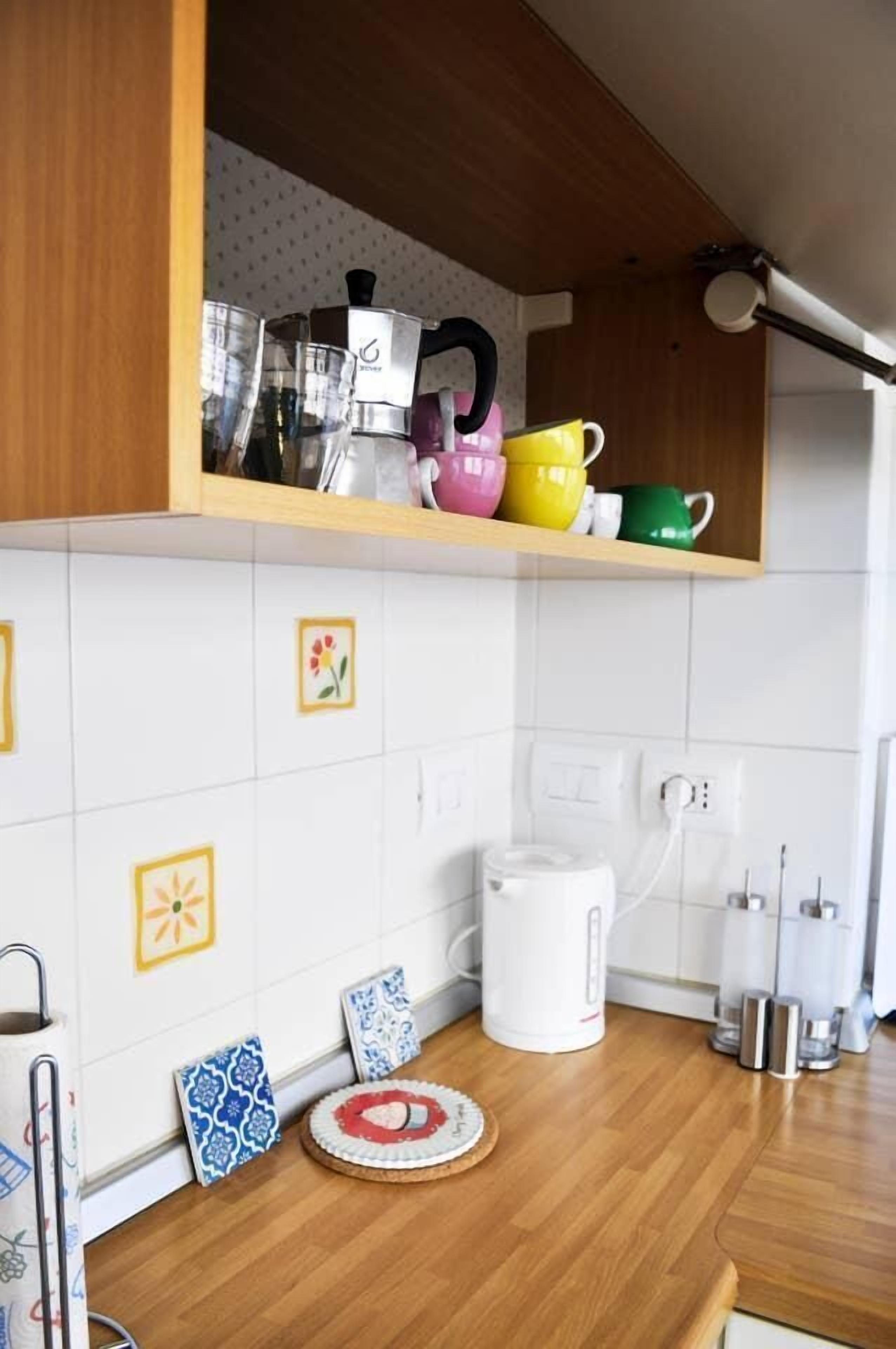 A kitchen with white tiles, a wooden countertop, a coffee maker, and colorful mugs on a shelf.