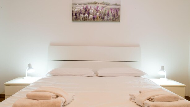 A neatly made bed with two folded towels, a white headboard, and a painting of flowers on the wall. - Casa Luisa (San Lazzaro di Savena)