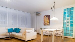 A modern living room with a white sofa, a dining table with clear chairs, and a large window with blue-tinted glass panels. - Casa Luisa (San Lazzaro di Savena)