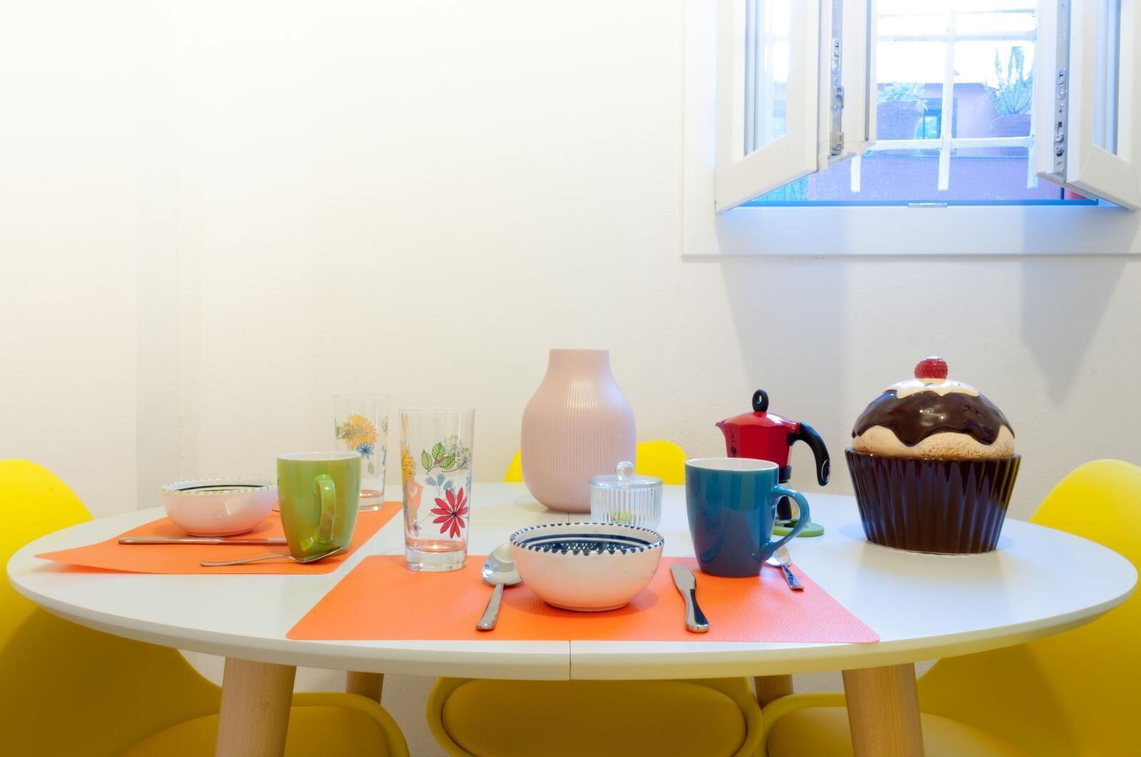 Une table ronde à la surface blanche, entourée de chaises jaunes. Sur la table, on trouve divers objets, notamment une tasse verte, une tasse bleue, un vase rose, une bouilloire rouge, un bol blanc, un bol bleu, une cuillère, une fourchette et un petit gâteau.
