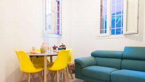 A dining area with a table and four yellow chairs, a teal sofa, and two windows with curtains. - Casa Greta (Bologna)