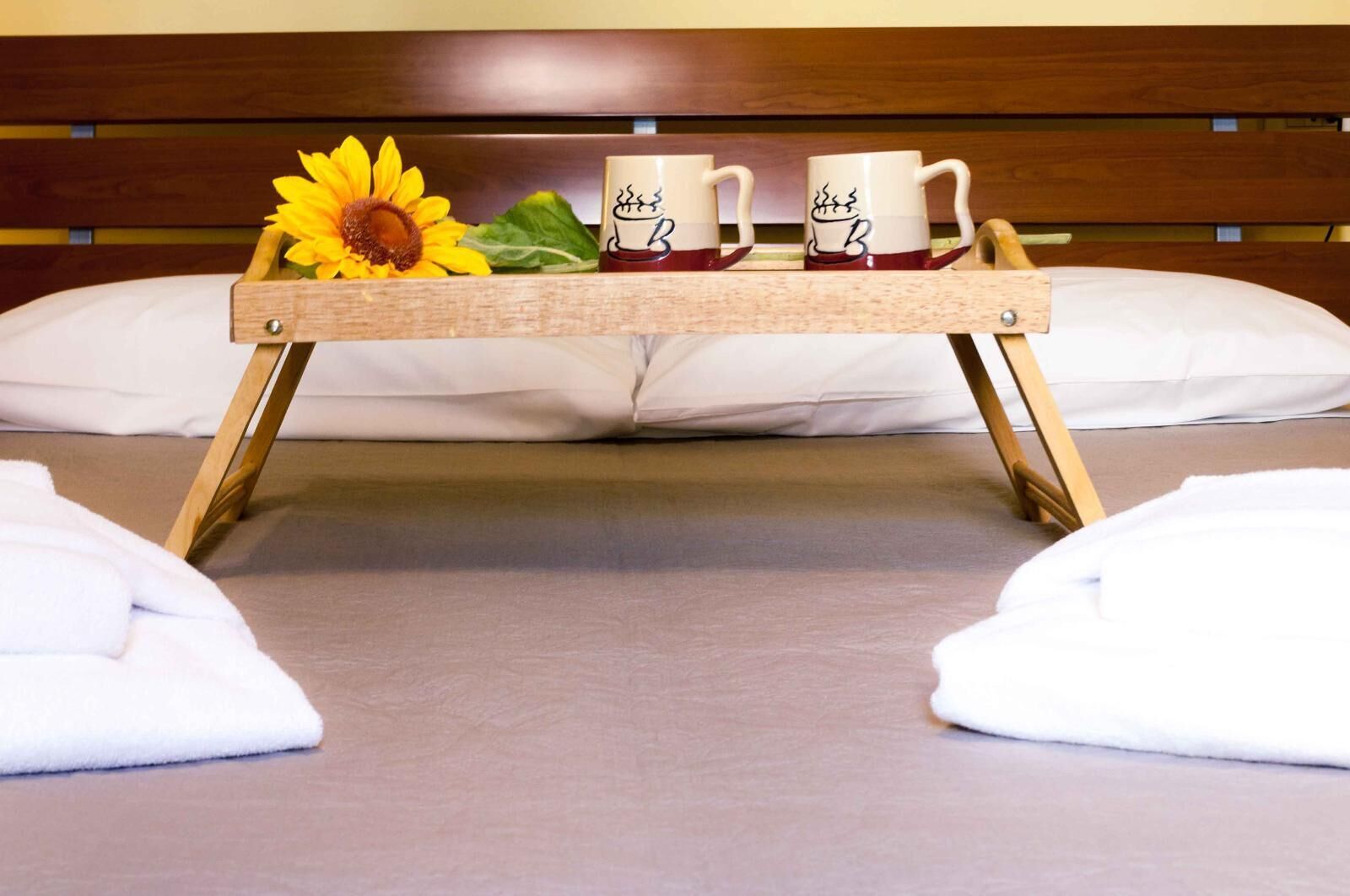 Een bed met een houten dienblad waarop drie mokken en een zonnebloem staan.