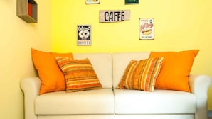 A white sofa with orange cushions against a yellow wall, decorated with signs for a donut shop, a coffee shop, and a hot dog stand. - Orange Flat - Fiera District (Bologna)