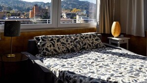 Een hotelkamer met een bed, een nachtkastje, een lamp en een uitzicht op een stadsgezicht door grote ramen.