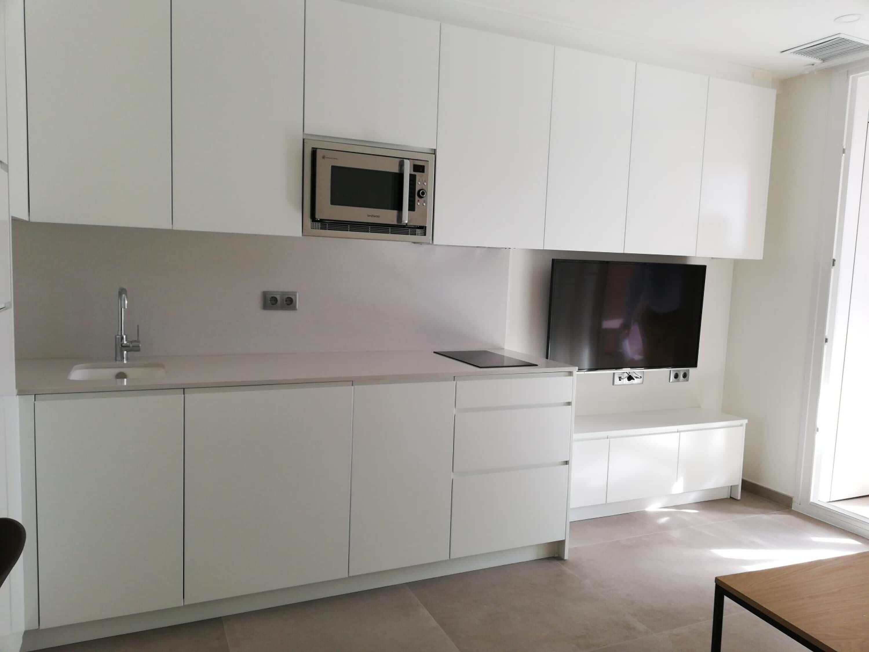 Una cocina moderna con armarios blancos, microondas de acero inoxidable, televisor de pantalla plana y un lavamanos.