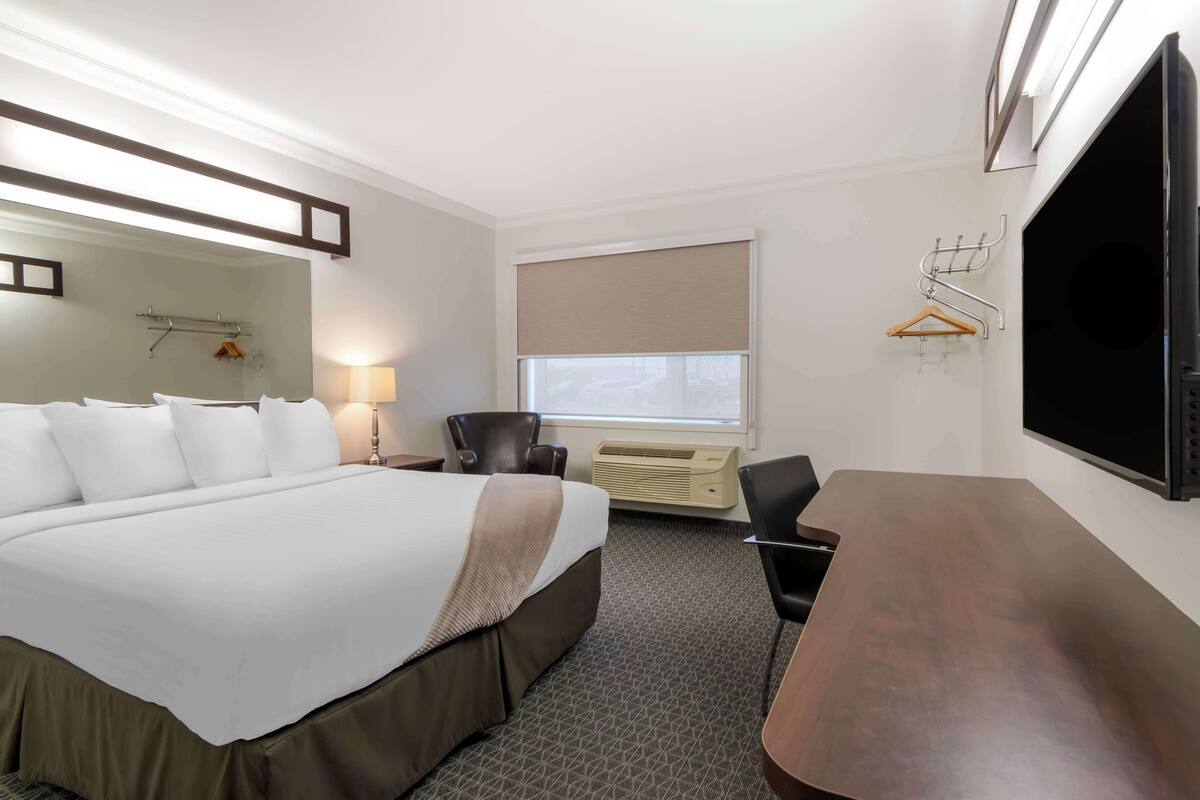 A hotel room with a large bed, a desk, a chair, a TV, a window with a blind, and a coat rack.