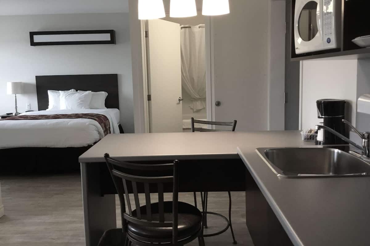 A compact hotel room with a bed, a small dining area with a chair, a table, a microwave, a coffee maker, and a sink.