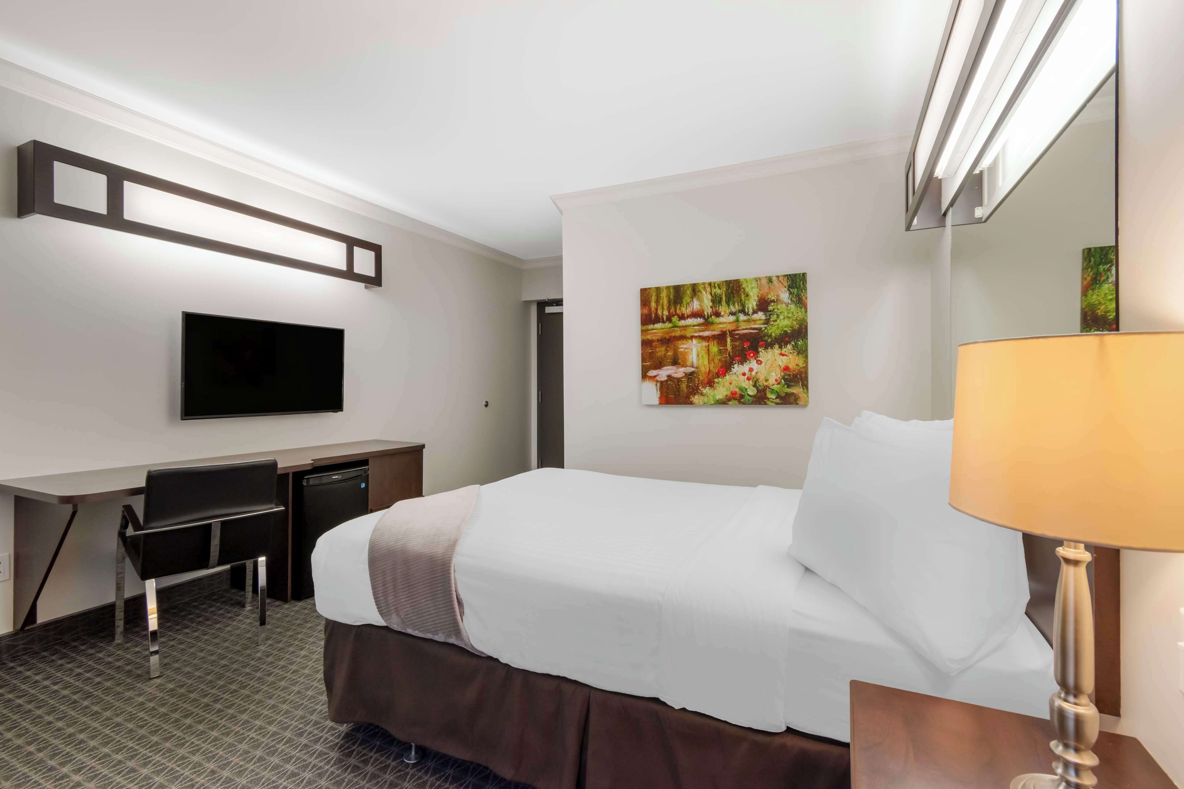 A hotel room with a bed, a desk, a chair, a TV, a painting, and a lamp.