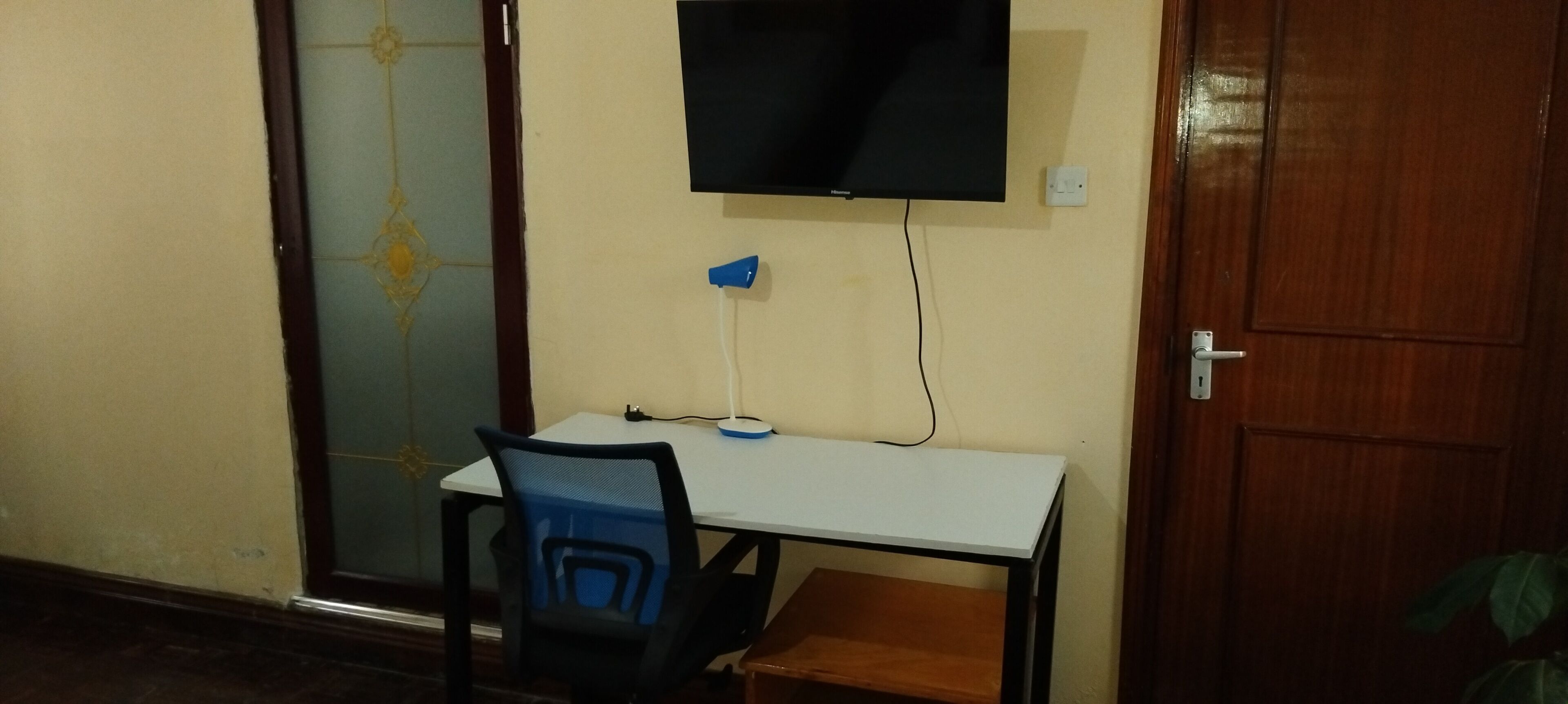Una habitación con un escritorio blanco, una silla azul, un televisor montado en la pared y una puerta de madera.