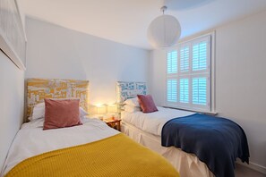 A room with two beds, a window with blinds, a hanging light fixture, and a wall with patterned wallpaper. - Charming, period cottage located just off the vibrant  high street of Deal with a gorgeous garden! (Deal)