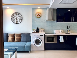 Un espace de vie compact comprenant un canapé, un lave-linge, un four à micro-ondes et un petit coin cuisine avec un évier et des placards.