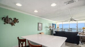 A kitchen with a white countertop, two wooden chairs, a ceiling fan, and a view of the ocean through large windows. - 0303 Waters Edge Resort 1 Bedroom Condo by RedAwning (Murrells Inlet)