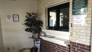 Uma área de recepção de hotel com parede de tijolos, uma janela, uma planta, balcão de check-in e diversos avisos, incluindo horários de check-out e opções de pagamento.