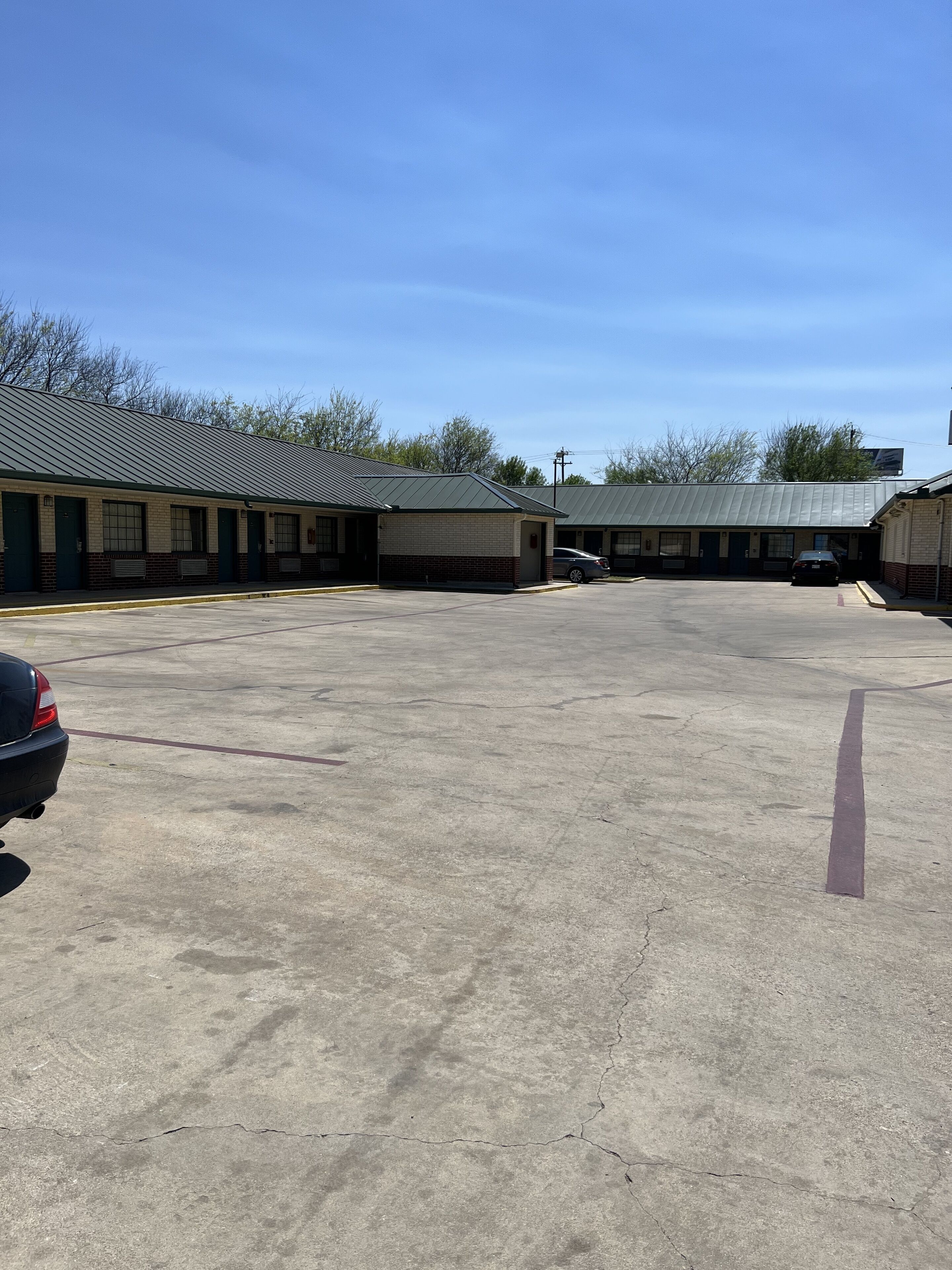 Um estacionamento com uma fileira de edifícios, incluindo um motel com toldo verde e uma construção térrea com telhado de metal.