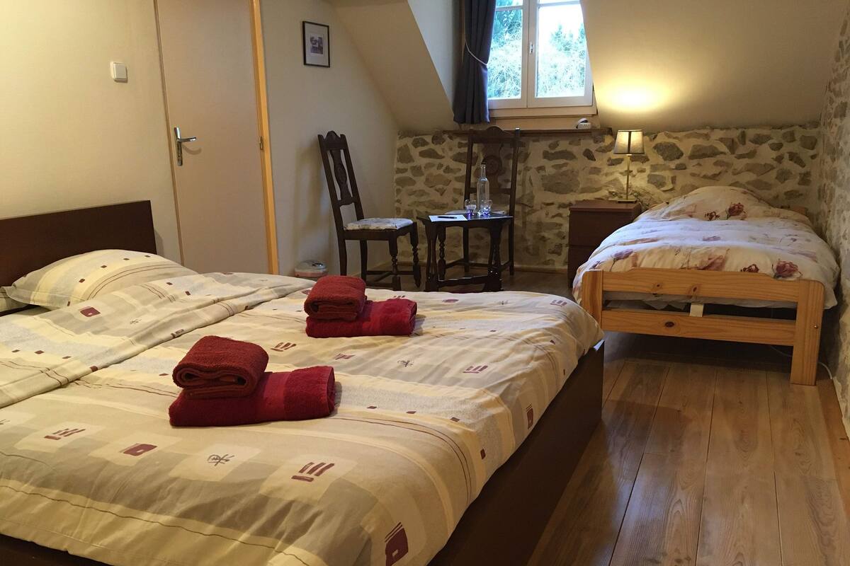 A bedroom with two beds, a wooden floor, a stone wall, a window with curtains, a chair, a small table, and a lamp.