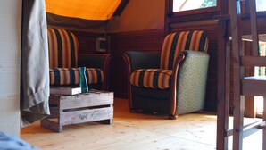 Een gezellige kamer met een houten vloer, twee gestreepte fauteuils, een klein houten tafeltje met een vaas, en een raam met uitzicht op groen.