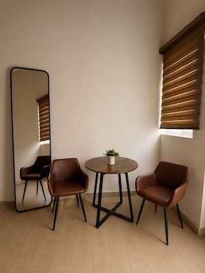 A room with a round table, two chairs, a potted plant, a large mirror, and a window with blinds. - Hotel Queretaro (Querétaro)