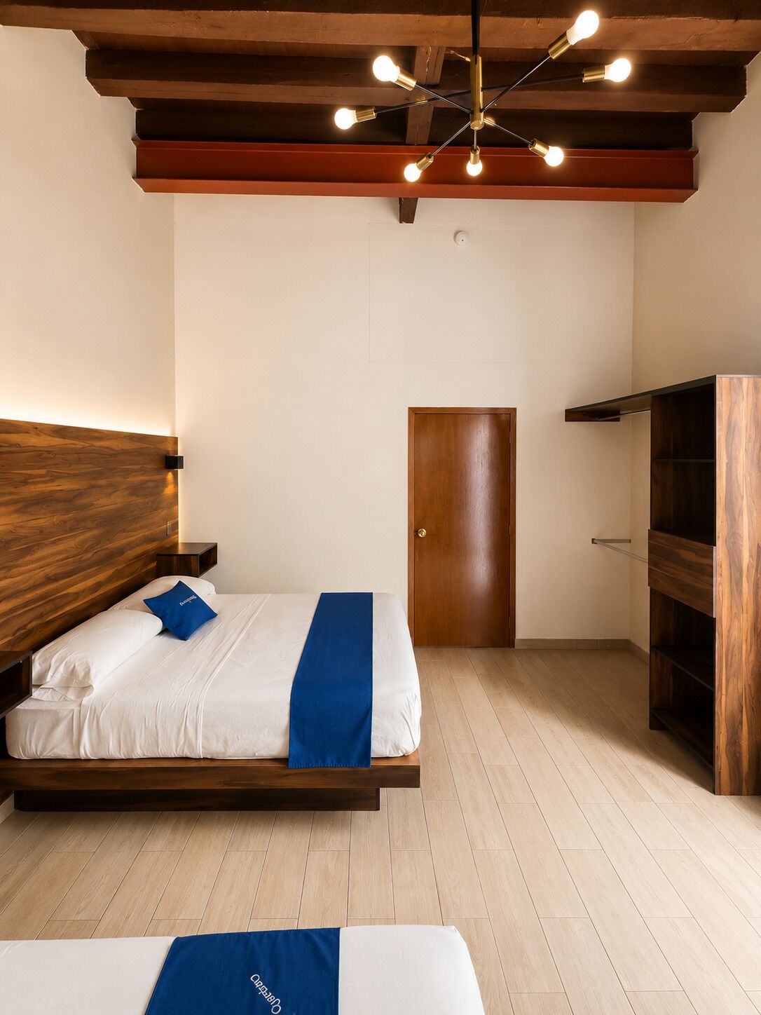 Habitación de hotel con cabecera de madera, cama con ropa blanca y detalles azules, armario de madera y lámpara en el techo.