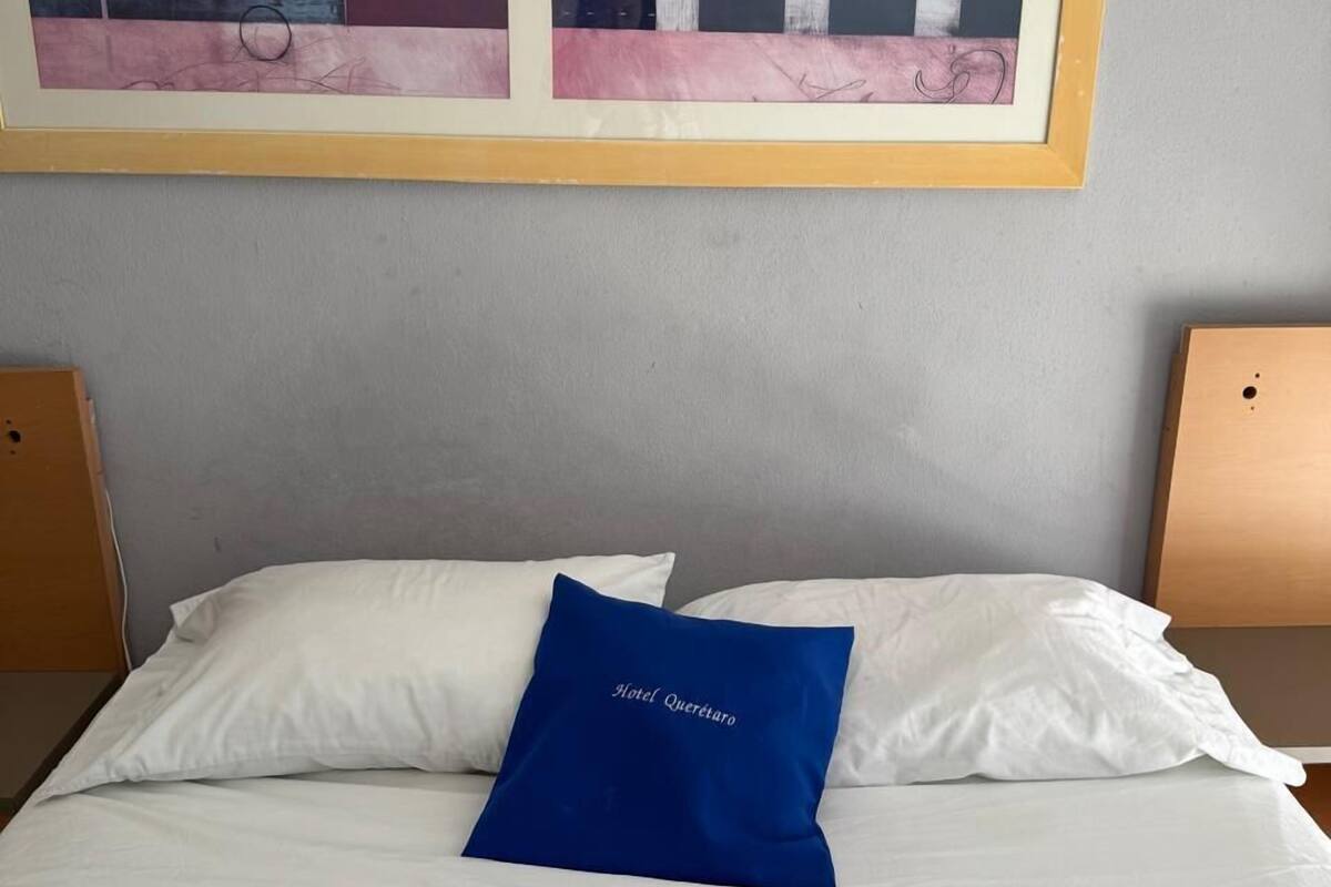 Une chambre d’hôtel avec un lit, du linge de lit blanc, un oreiller bleu orné du texte « Hotel Querétaro », et un tableau abstrait encadré au-dessus du lit.