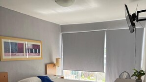 Una habitación de hotel con cama, escritorio, silla, basurero, luz de techo, ventana con persianas y un cuadro en la pared.