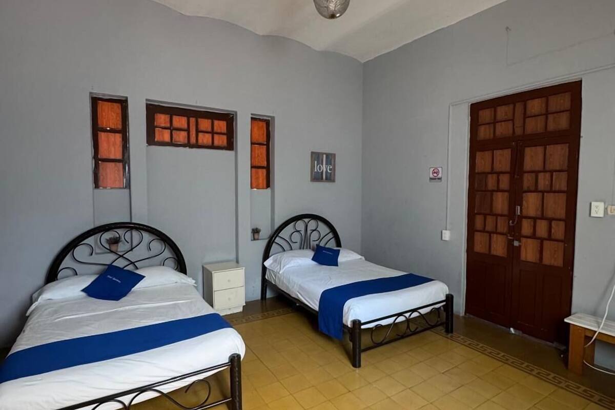 Une chambre avec deux lits simples, une porte en bois, une petite armoire blanche et un luminaire au plafond.