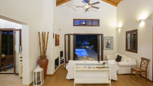 A spacious living area with a high ceiling, a fireplace, a sofa, and a view of a swimming pool at night.