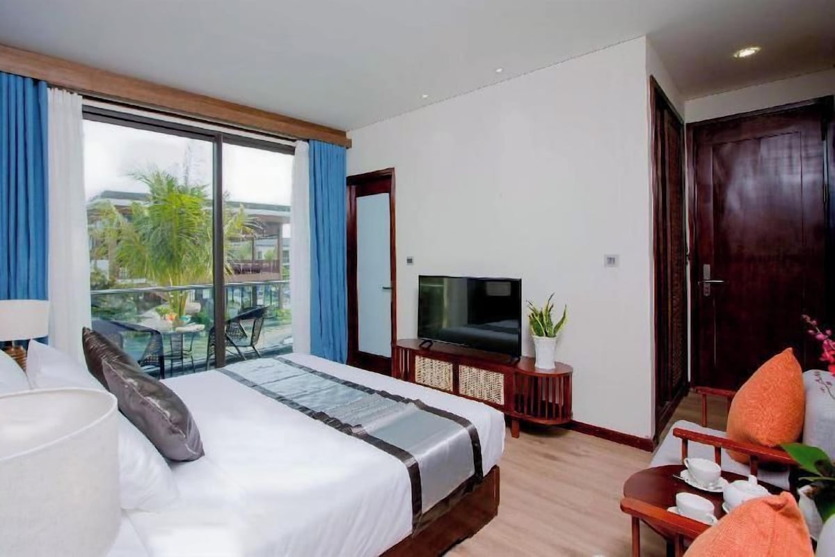 Une chambre d’hôtel avec un grand lit, une télévision, une petite table avec une plante et un balcon donnant sur des palmiers.