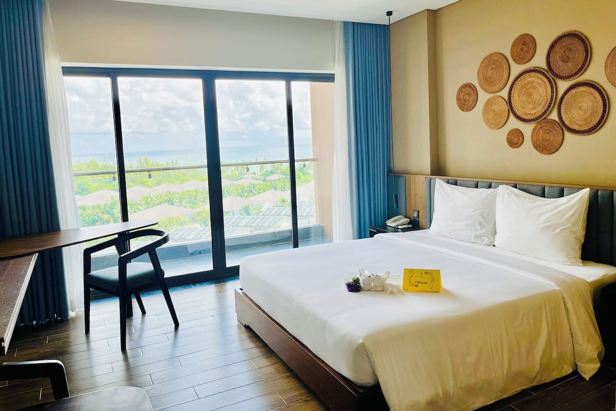 Une chambre d’hôtel avec un grand lit, un bureau et une chaise. La fenêtre offre une vue sur la verdure.