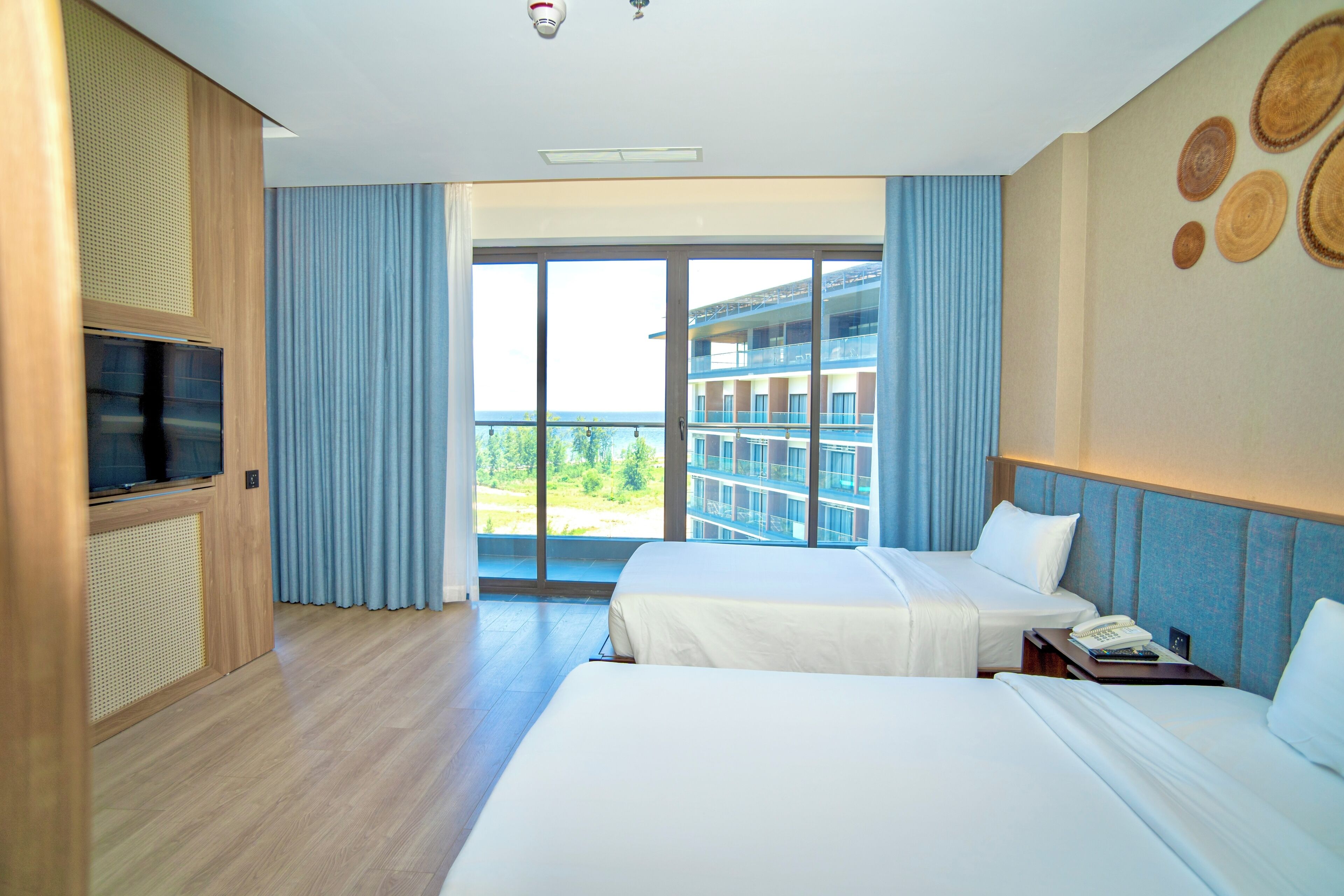 A hotel room with two beds, a TV, and a large window with a view of a building and greenery.