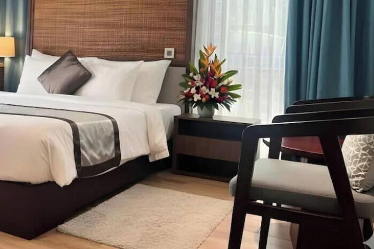 Un lit bien fait, avec une tête de lit sombre, une table de chevet sur laquelle se trouve une lampe, et une chaise agrémentée d’un coussin.