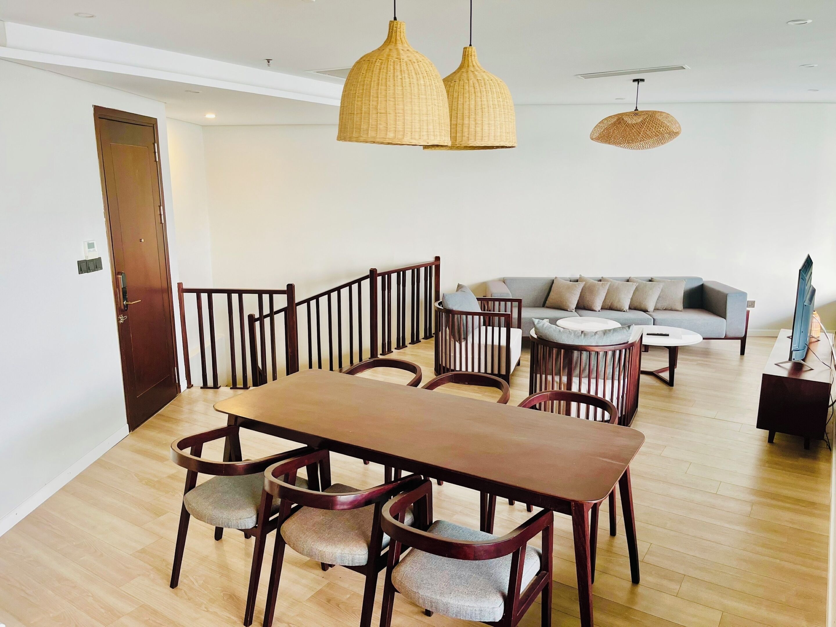 A modern living room with a wooden dining table, cushioned chairs, a grey sofa, and two hanging wicker lamps.