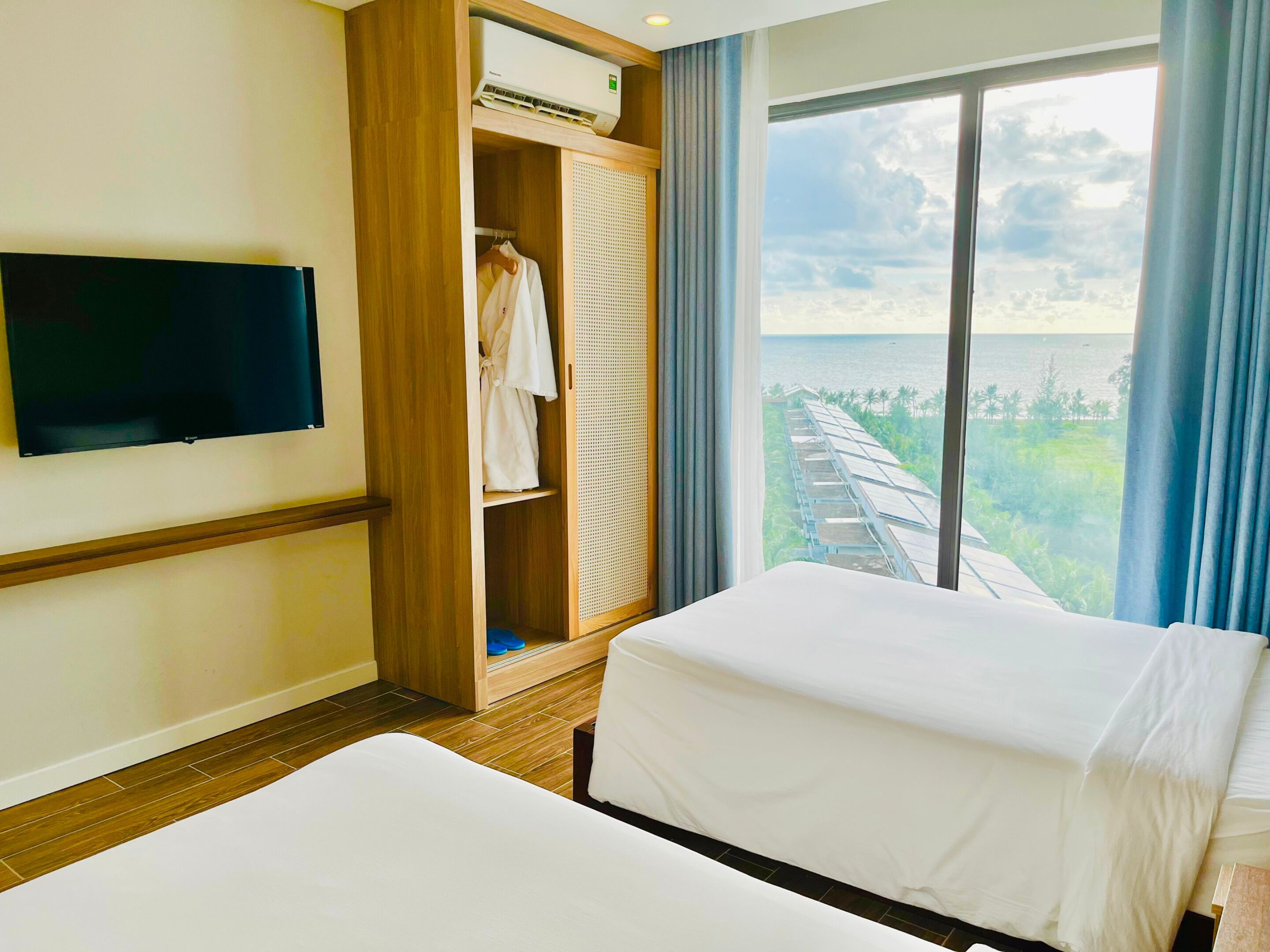 Habitación de hotel con cama, televisor, armario y una ventana grande con vista al mar y vegetación.