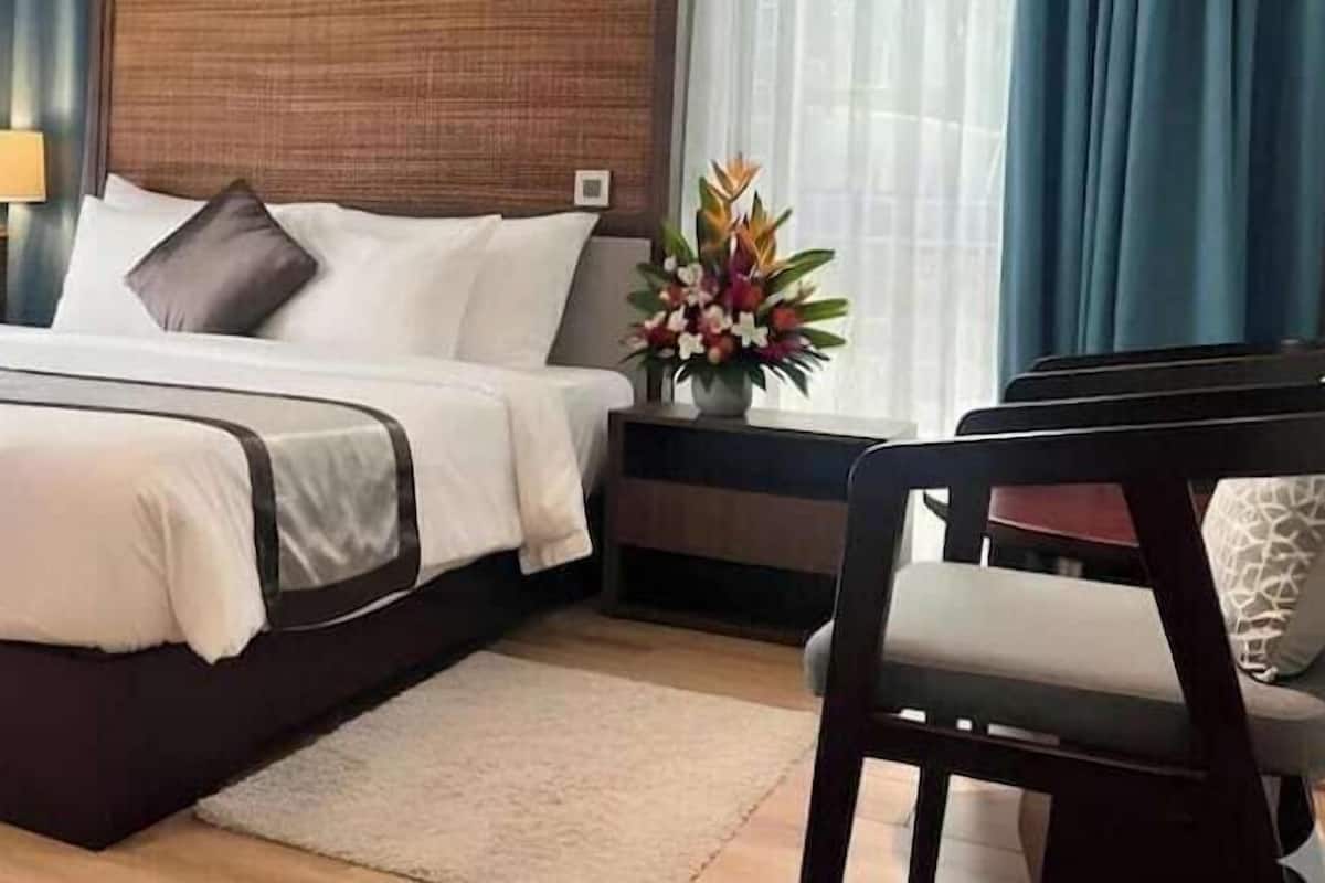 Une chambre d’hôtel avec un lit, une table de chevet, une lampe, une chaise et un vase de fleurs.