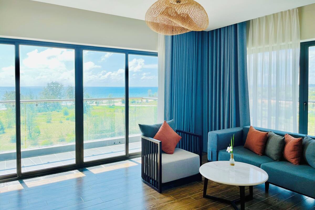 Une pièce spacieuse avec de grandes fenêtres donnant sur la mer et la verdure. On y trouve un canapé bleu, une petite table et un luminaire suspendu.