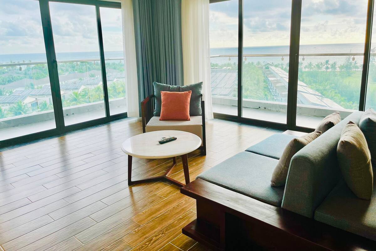 Une pièce spacieuse dotée d’une grande fenêtre offrant une vue sur la mer et la verdure. On y trouve un canapé, une petite table ronde et un fauteuil avec des coussins.
