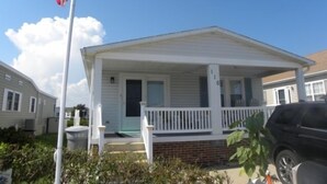 Een gelijkvloers huis met een veranda, een vlaggenmast met een Amerikaanse vlag, en een fundering van baksteen.
