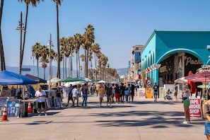 A bustling street with palm trees, market stalls, and a vape shop. - Amber Tide - Bright 1BR Marina Gem Ocean View (Marina Del Rey)