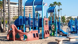 Ein Strandspielplatz mit Spielgeräten, darunter Rutschen und Kletterbereiche, umgeben von Palmen und einem Hotel im Hintergrund.