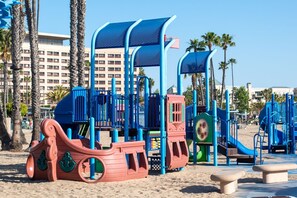 A beach playground with play structures, including slides and climbing areas, surrounded by palm trees and a hotel in the background.