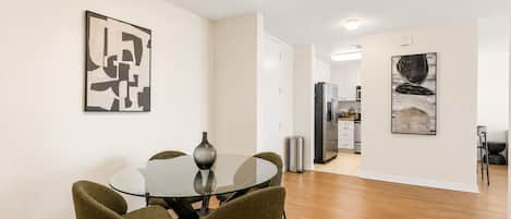 A modern dining area with a glass table and four chairs, a kitchen in the background, and abstract art on the walls.