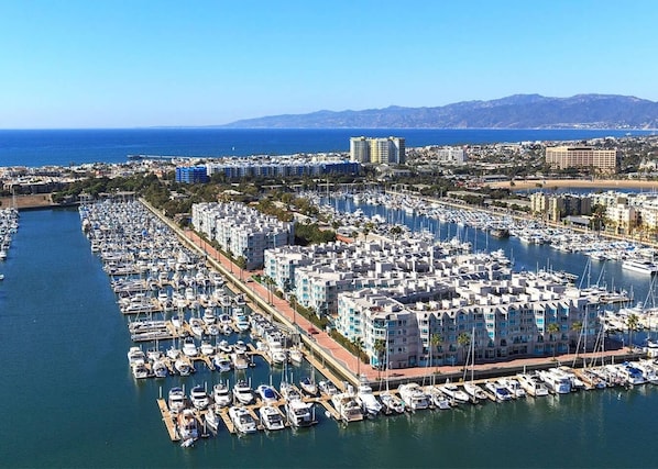 A marina with numerous boats docked, surrounded by residential buildings and a clear view of the sea and distant mountains. - Amber Tide - Bright 1BR Marina Gem Ocean View (Marina Del Rey)