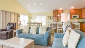 A spacious living room with a blue sofa, a brown armchair, and a white coffee table. There is a dining area with a table and chairs, and a kitchen with wooden cabinets and appliances. - Montego Bay Community Canal Front  wrap around porch, boat tie up, Pools, Tennis N. Ocean City MD! (Ocean City)