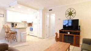A living room with a wooden coffee table, a TV stand with a flat-screen TV, and a green armchair. - Ocean Block Condo, porch, 1st floor, steps to beach, updated, full kitchen, lg dining table, wifi! (Ocean City)