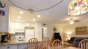 A compact kitchen with white cabinets, a dining table with chairs, a television, and a decorative ceiling light. - Ocean Block Condo, porch, 1st floor, steps to beach, updated, full kitchen, lg dining table, wifi! (Ocean City)