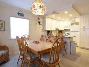 Uma sala de jantar com mesa e cadeiras de madeira, um lustre e uma cozinha ao fundo.