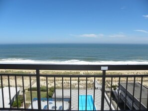 A balcony with a view of the ocean. There is a pool with lounge chairs and a building in the background.