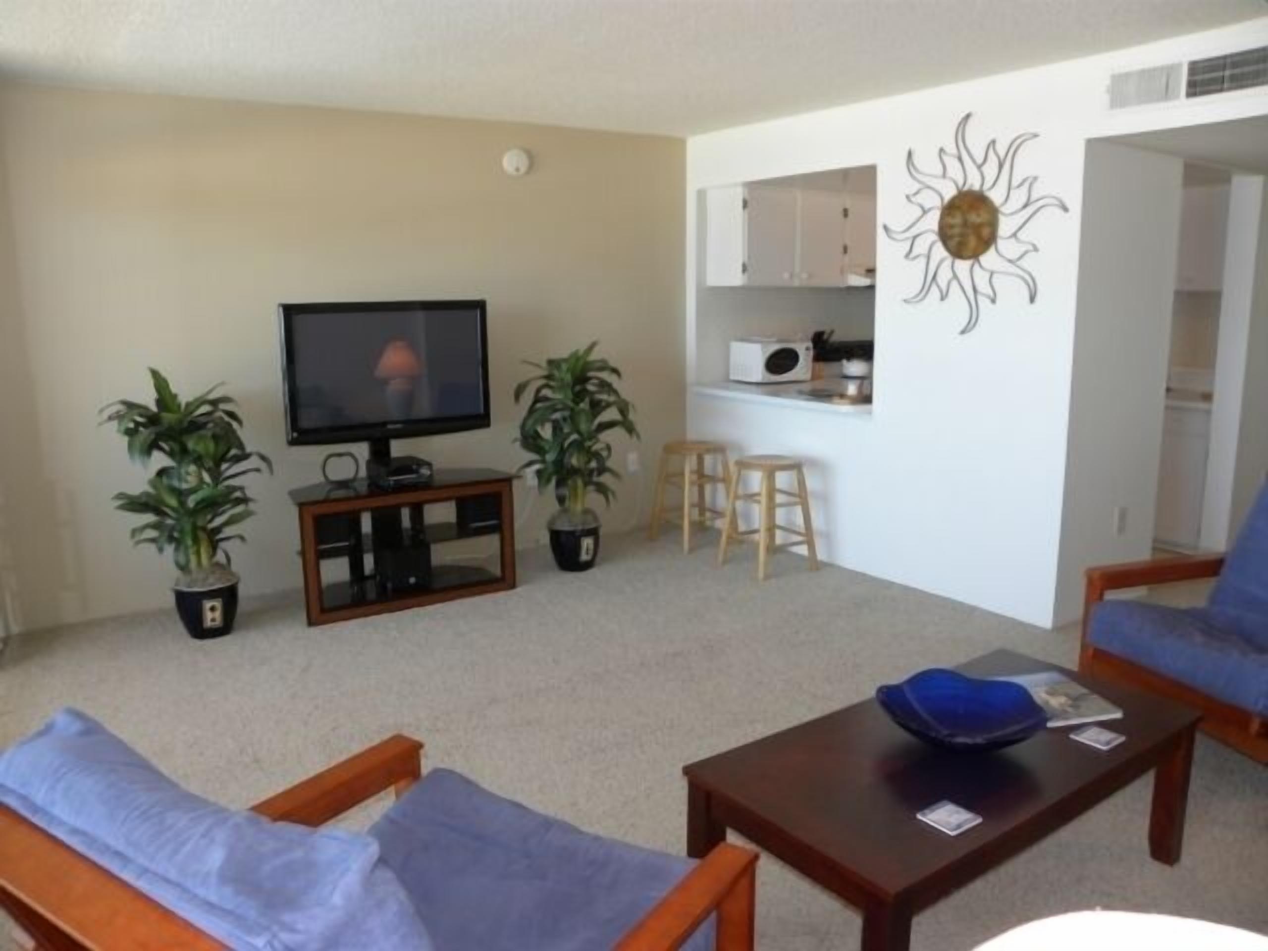 A living room with a TV, two potted plants, a blue sofa, a wooden coffee table, and a kitchen area with stools.