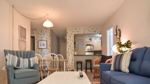 A living room with a blue sofa, a white coffee table, and a blue armchair. There is a kitchen in the background with white cabinets and a hanging light fixture. - Ocean Front Building w/ side views of Ocean and Beach, lifeguarded pool, Game Room, Gym, Tennis (Ocean City)